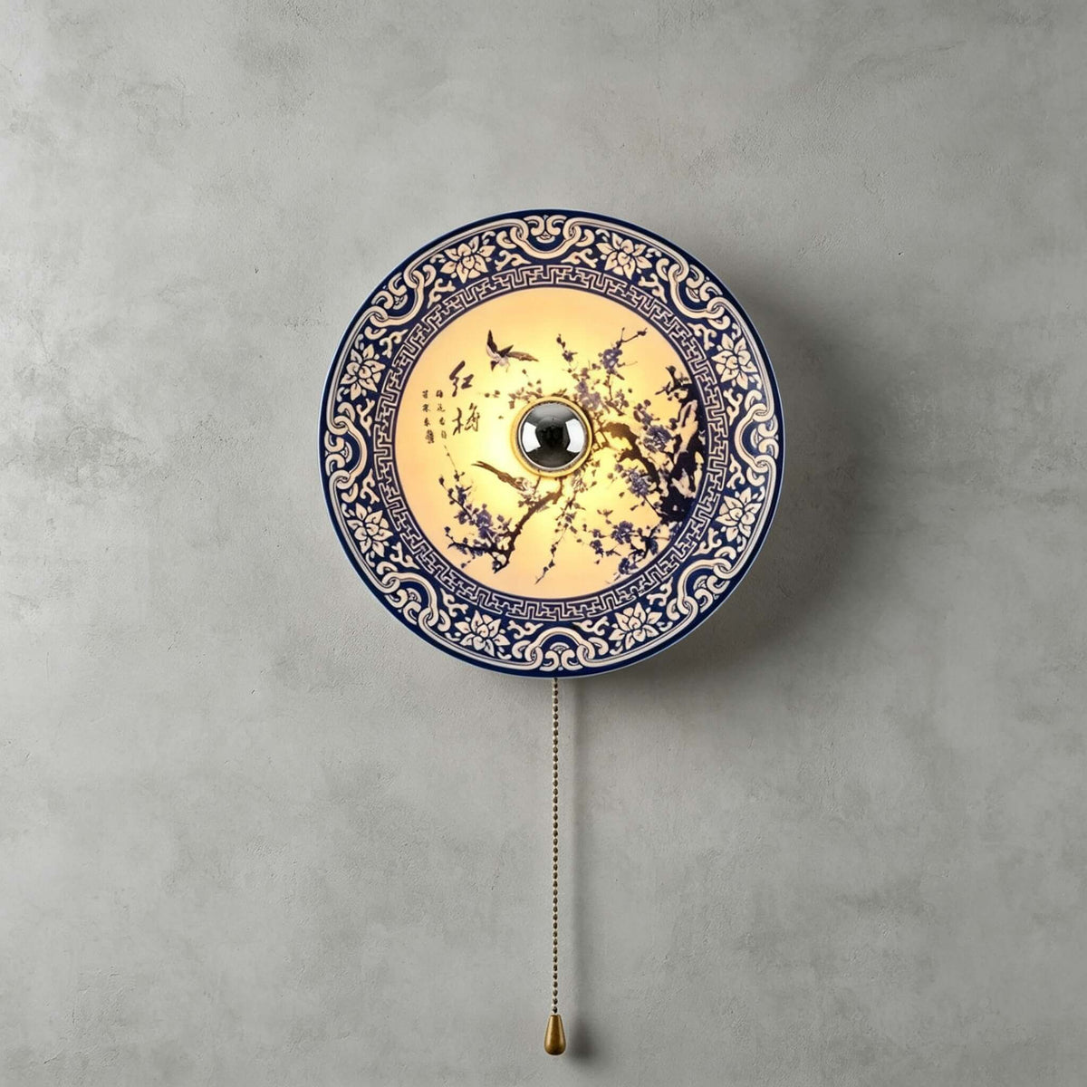 Lit Clérachic wall light with a blossom-branch design and a patterned blue border, its pull chain hanging on a grey wall.