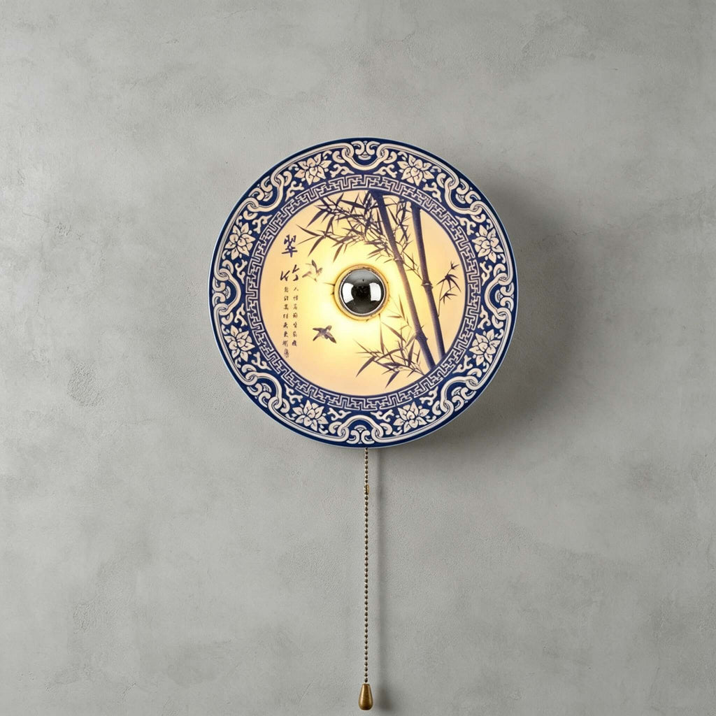 Clérachic wall light with a bamboo and bird design inside a patterned blue border, a brass chain below, on a grey wall.