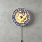 Clérachic wall light with a bamboo and bird design inside a patterned blue border, a brass chain below, on a grey wall.