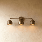 Clérachic three-light bar sconce in vintage brass with dome shades, mounted on a plaster wall in studio light.