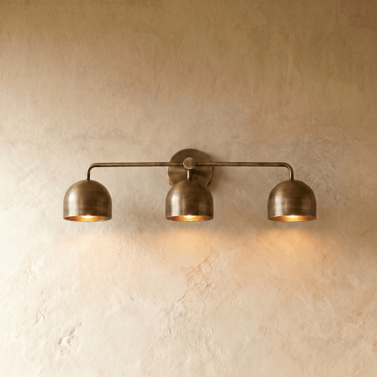Clérachic three-light bar sconce in vintage brass with dome shades, mounted on a plaster wall in studio light.