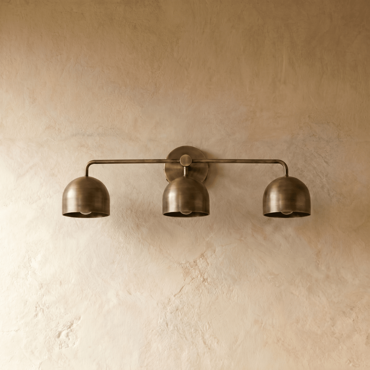 Clérachic three-light bar sconce in vintage brass, shown from a slightly closer studio angle on a plaster wall.