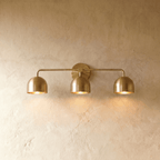 Clérachic three-light bar sconce in polished brass with dome shades, mounted on a plaster wall in studio light.