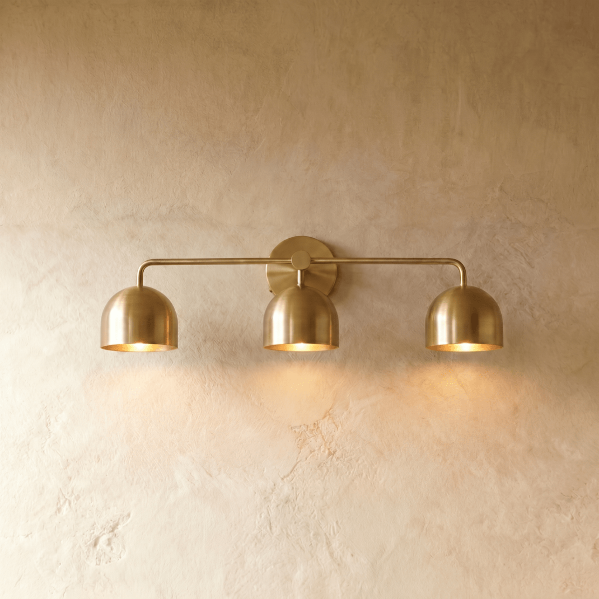 Clérachic three-light bar sconce in polished brass with dome shades, mounted on a plaster wall in studio light.