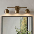 Clérachic brass three-light bar sconce lit above a black-framed mirror with a leafy plant in reflection.