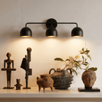Clérachic matte black three-light bar sconce lit above a console displaying carved wooden figures and pottery.