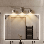 Clérachic chrome three-light bar sconce lit above a backlit mirror beside a black towel warmer in a bathroom.