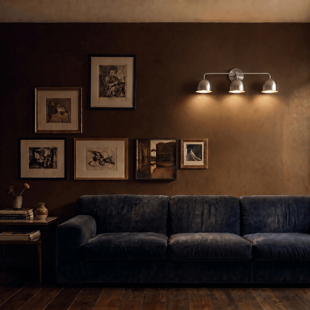 Clérachic chrome three-light bar sconce lit above a navy sofa beside a gallery wall of framed prints.