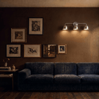 Clérachic chrome three-light bar sconce lit above a navy sofa beside a gallery wall of framed prints.