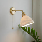 Lit Clérachic wall light with a ribbed white ceramic shade on a brass arm, angled out from a grey wall beside a green fern.