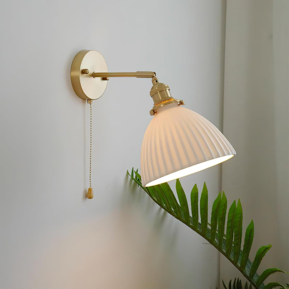 Lit Clérachic wall light with a ribbed white ceramic shade on a brass arm, angled out from a grey wall beside a green fern.