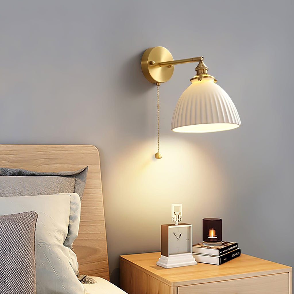 Pleated ceramic shade of this Clérachic wall light glowing above a pale wood bedside table, with a clock and candle beside the bed.
