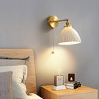 Pleated ceramic shade of this Clérachic wall light glowing above a pale wood bedside table, with a clock and candle beside the bed.