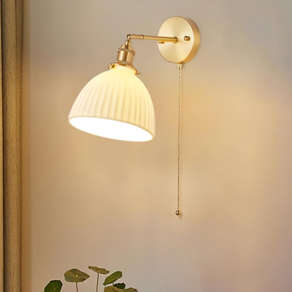 A Clérachic brass wall light with a fluted ceramic shade casting warm light on a soft peach wall, a small plant at the bottom.