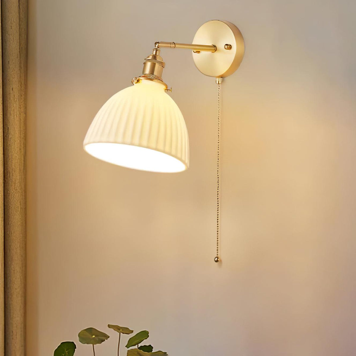 A Clérachic brass wall light with a fluted ceramic shade casting warm light on a soft peach wall, a small plant at the bottom.