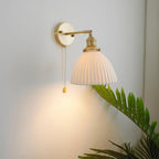 Brass-armed Clérachic wall light with a pleated white shade, lit and hanging low on a grey wall with a fern leaf nearby.
