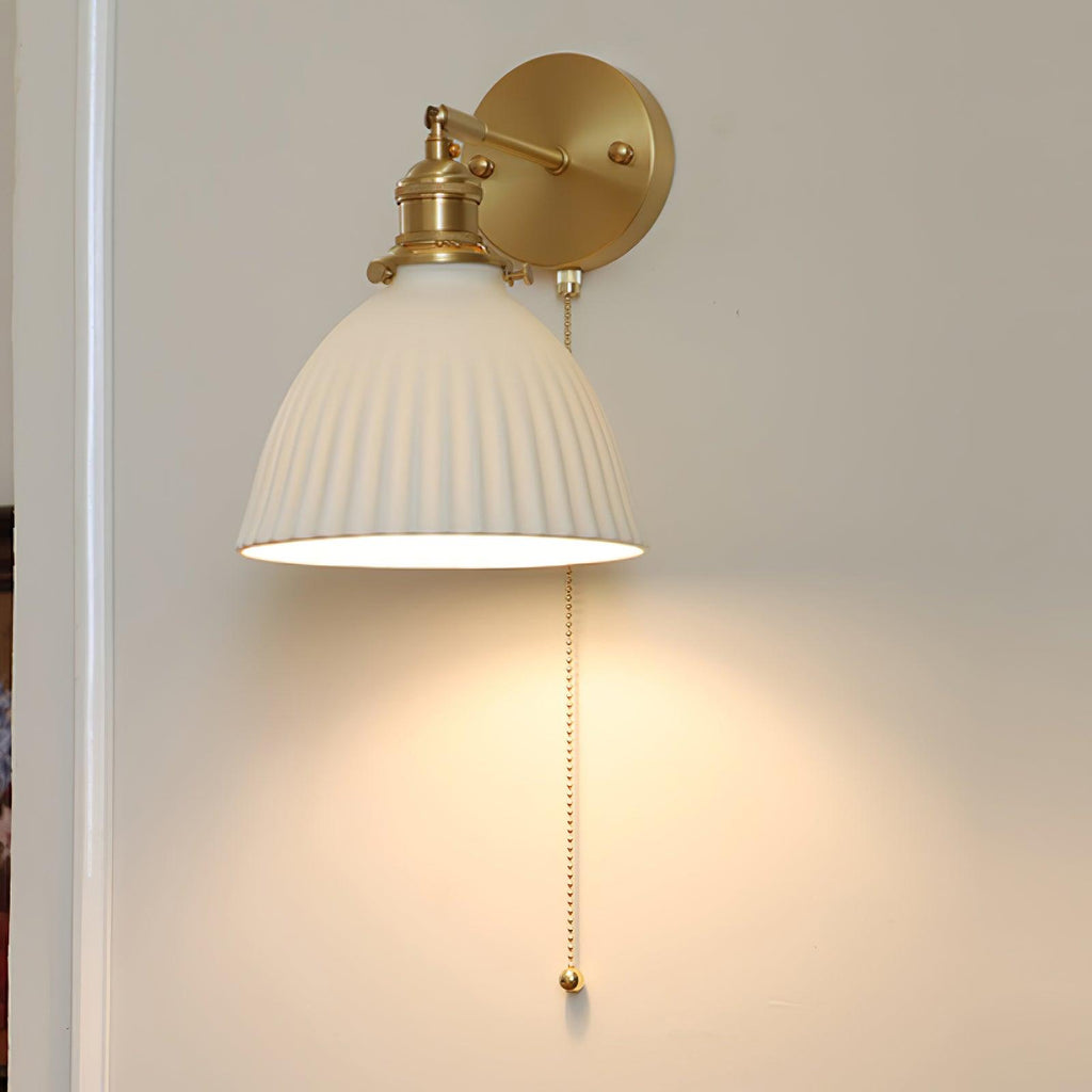 Close front view of a lit Clérachic wall light, its wide pleated ceramic shade hung from a round brass backplate.