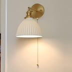 Close front view of a lit Clérachic wall light, its wide pleated ceramic shade hung from a round brass backplate.