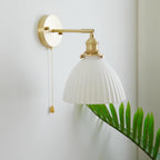 Unlit pleated white ceramic shade on a brass arm and round backplate, this Clérachic wall light shown against a pale wall with a fern.