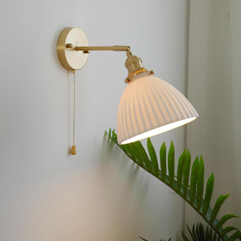 A Clérachic wall light with a tilted, pleated ceramic shade and a brass swing arm, glowing softly over a fern on a grey wall.