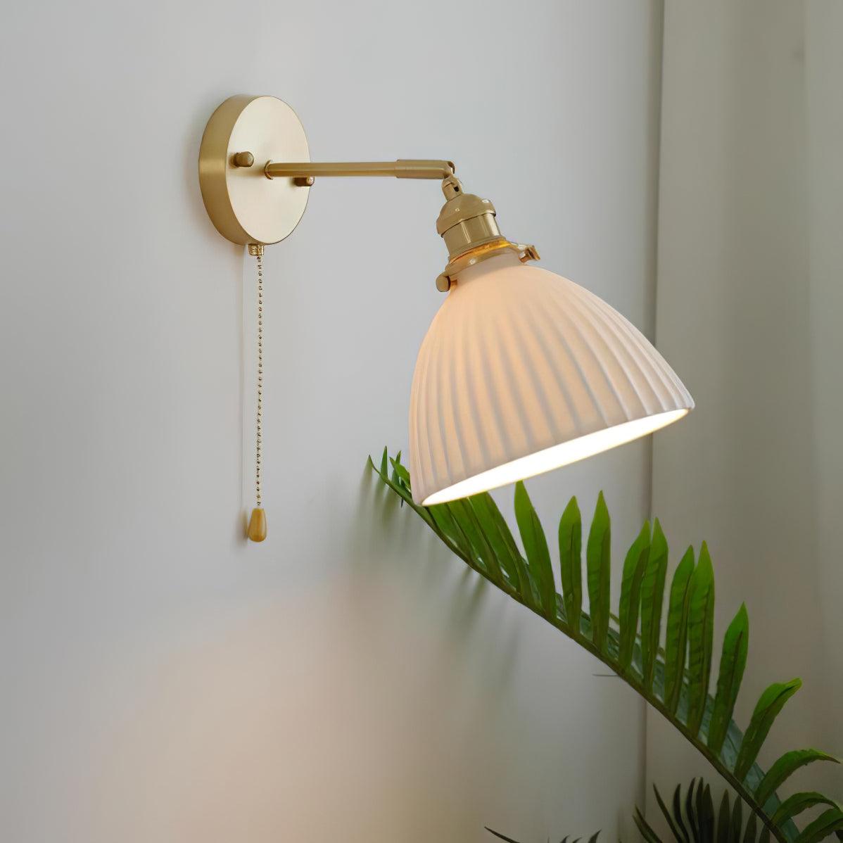 A Clérachic wall light with a tilted, pleated ceramic shade and a brass swing arm, glowing softly over a fern on a grey wall.