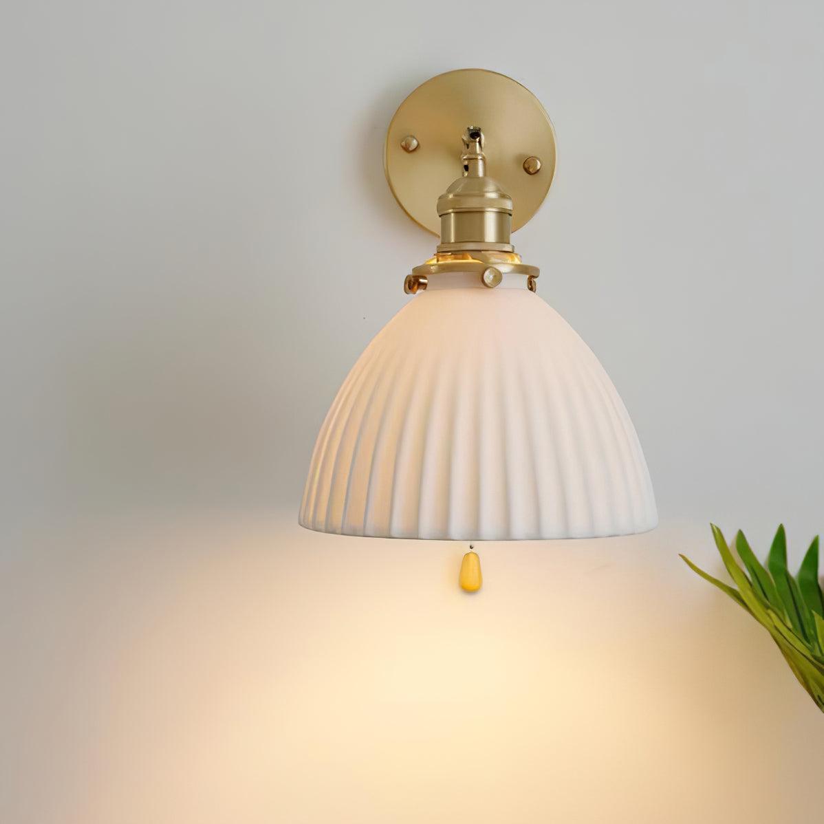 Front view of the brass socket, round backplate and bell-shaped pleated shade of this Clérachic wall light, lit warm.