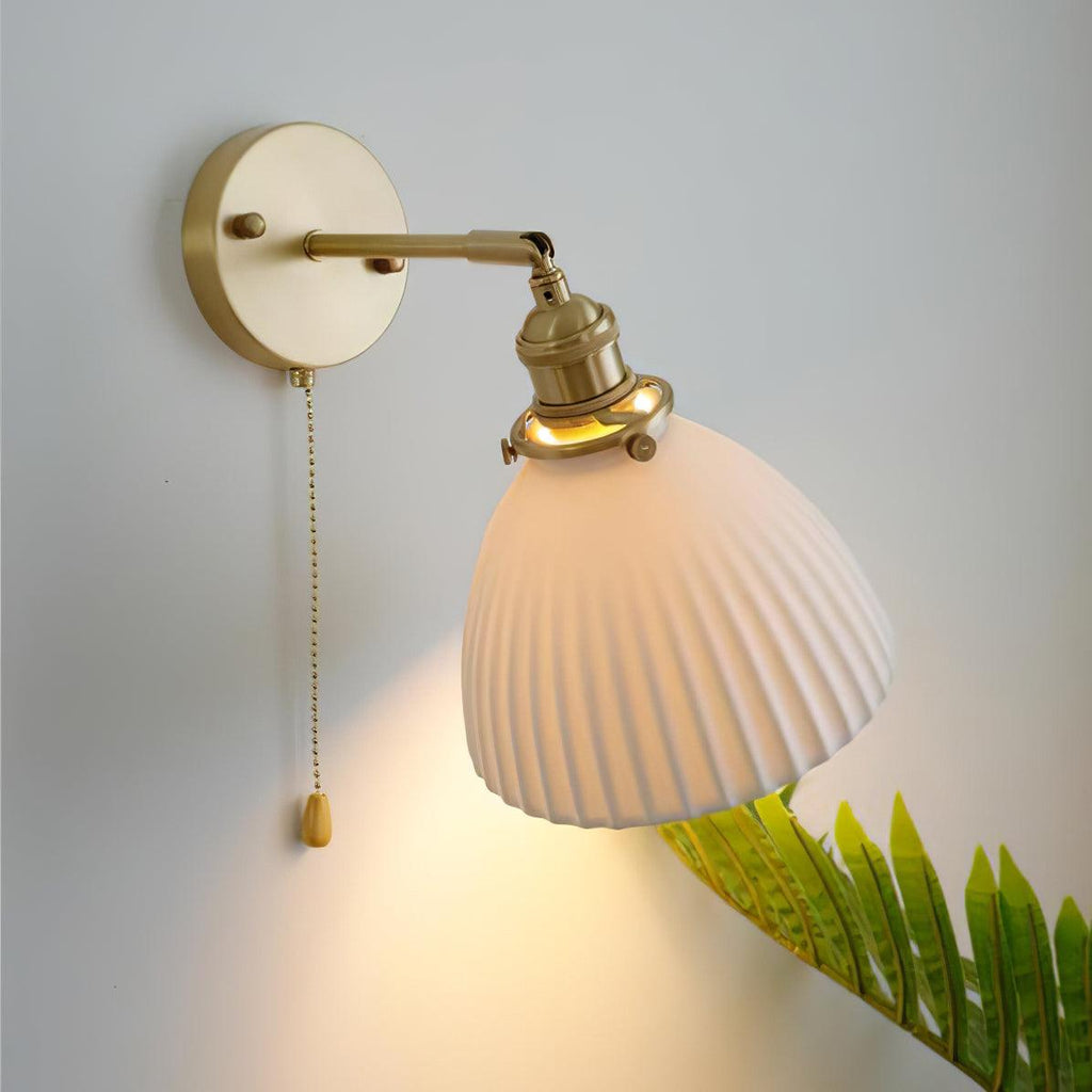 Pleated ceramic shade of this Clérachic wall light glowing from within, held out on a curved brass arm with a fern beside it.