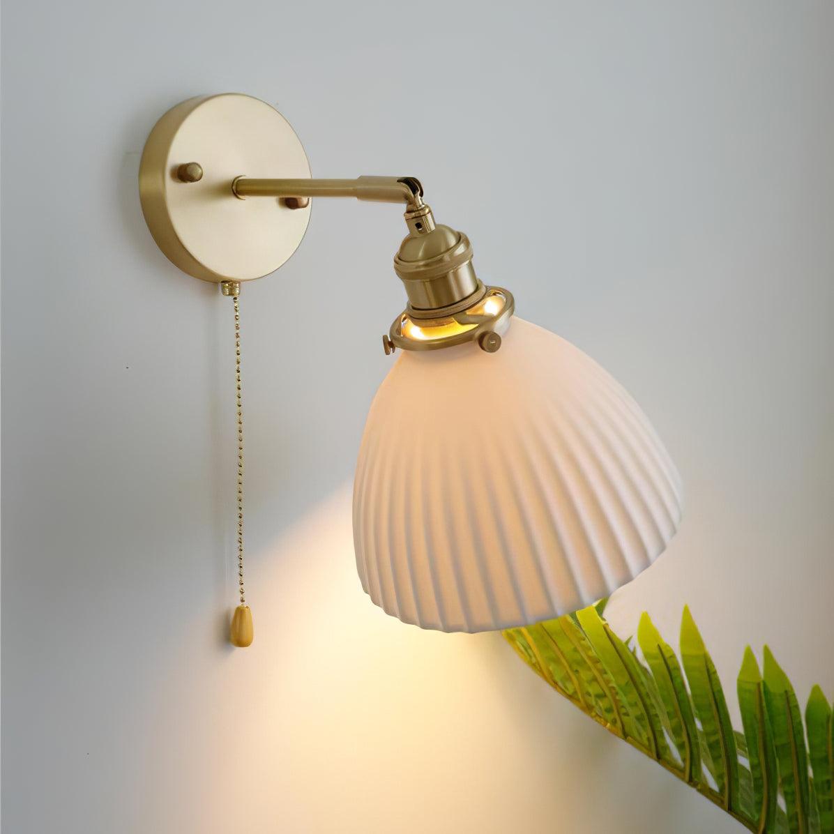 Pleated ceramic shade of this Clérachic wall light glowing from within, held out on a curved brass arm with a fern beside it.
