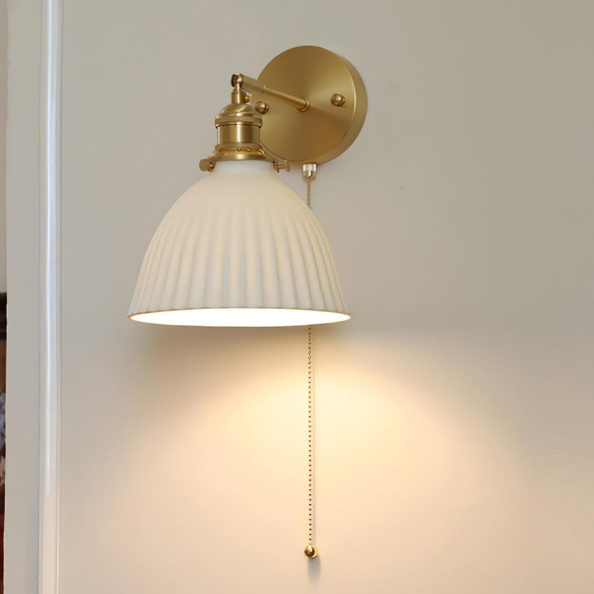 A Clérachic brass wall light with a wide, pleated white shade and pull chain, lit against a warm cream wall.