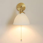 Straight-on view of the Clérachic wall light, its pleated ceramic bell shade under a brass fitting, lit on a cream wall.