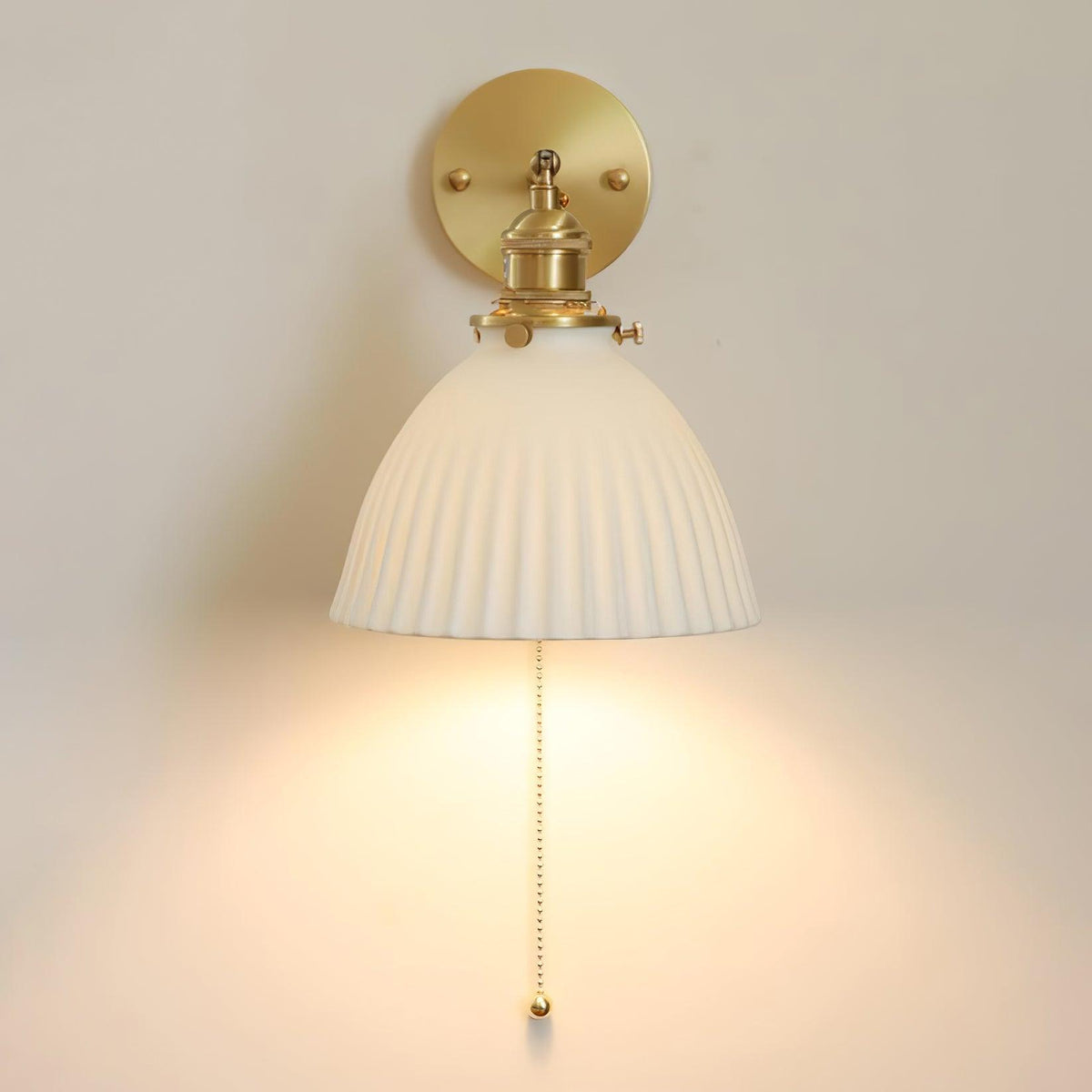 Straight-on view of the Clérachic wall light, its pleated ceramic bell shade under a brass fitting, lit on a cream wall.