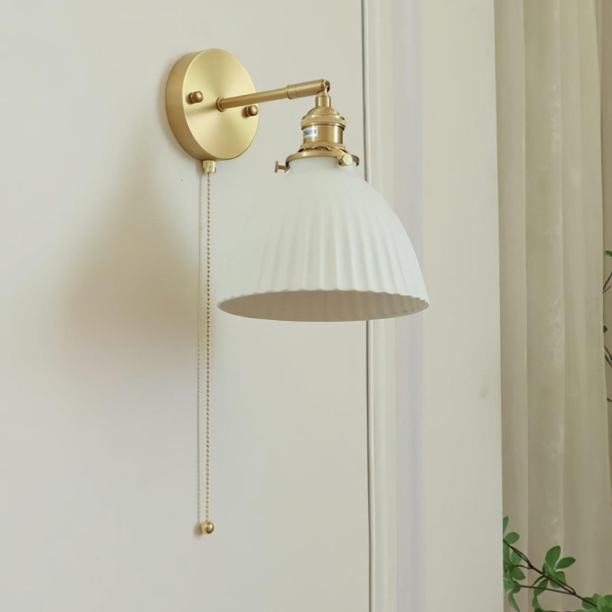 Clérachic wall light with a pleated ceramic shade tilted to show its inside, on a brass arm against a pale wall and curtain.