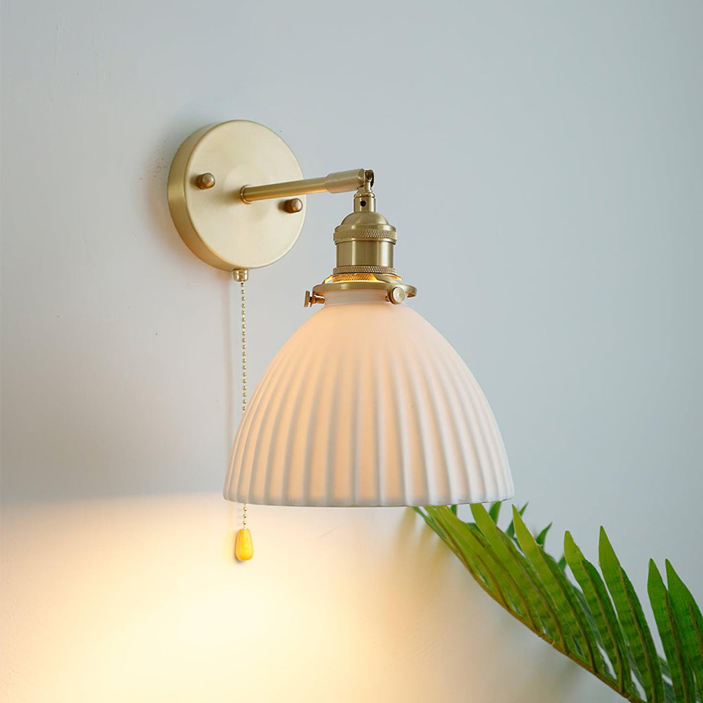 Lit Clérachic wall light with a ribbed ceramic shade glowing warm, a brass backplate above and a gold pull-chain pendant below.