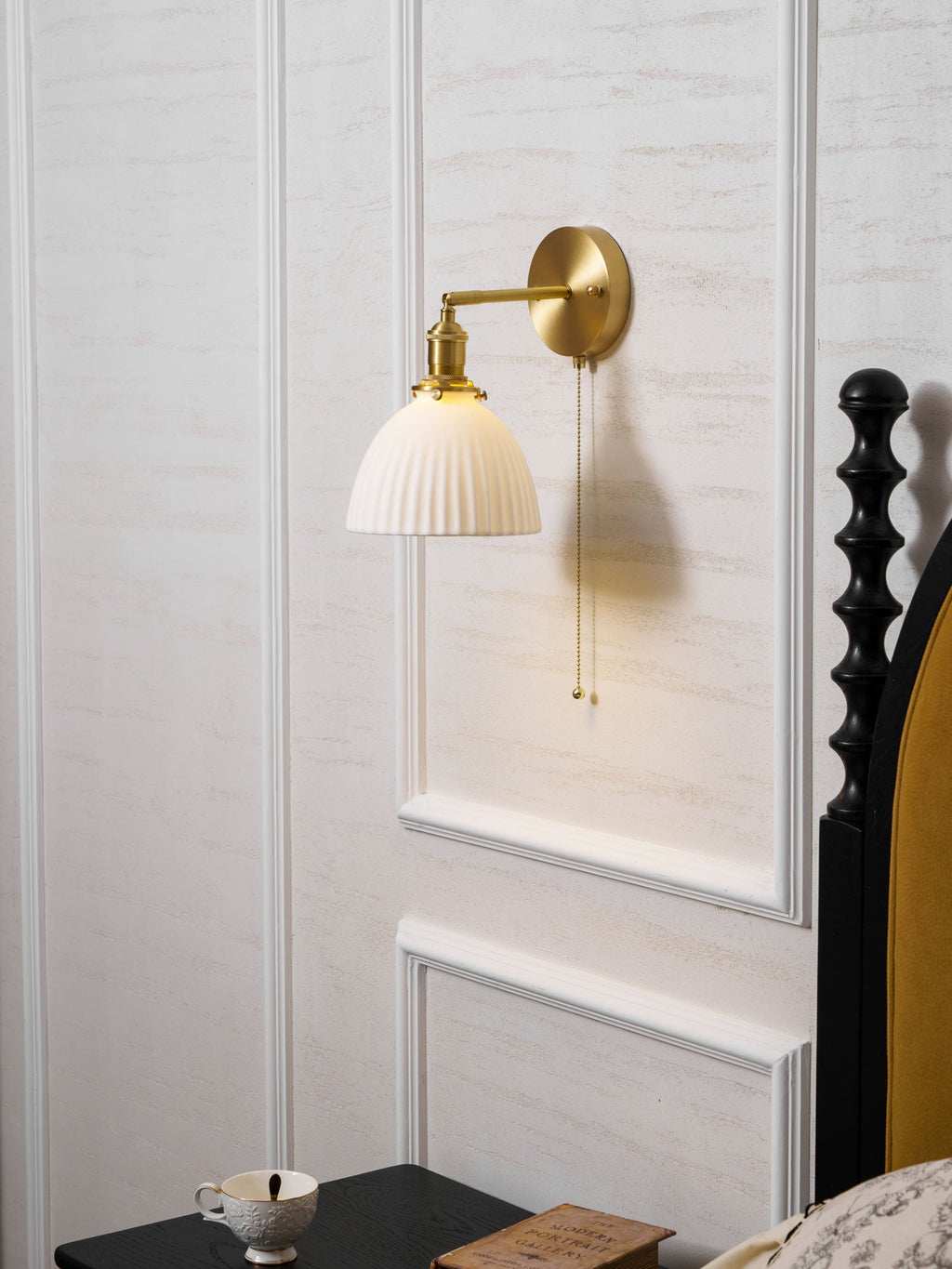 Clérachic wall light on a white panelled wall beside a black carved bedpost and a mustard headboard, its shade unlit.