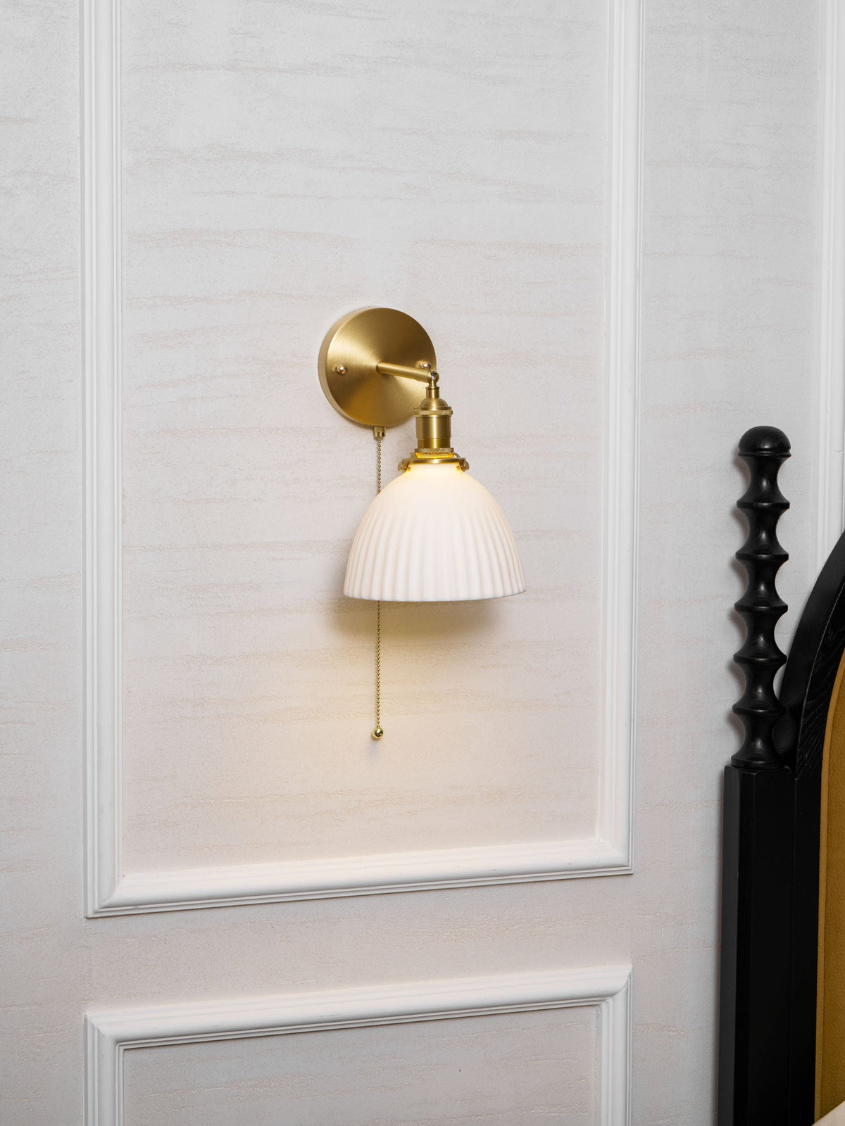 A Clérachic wall light with a pleated white shade glowing low on a panelled bedroom wall, close to a black bed frame.