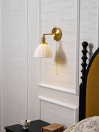 Clérachic wall light with its brass arm angled left from the backplate, over a dark bedside table with a cup and a small book.