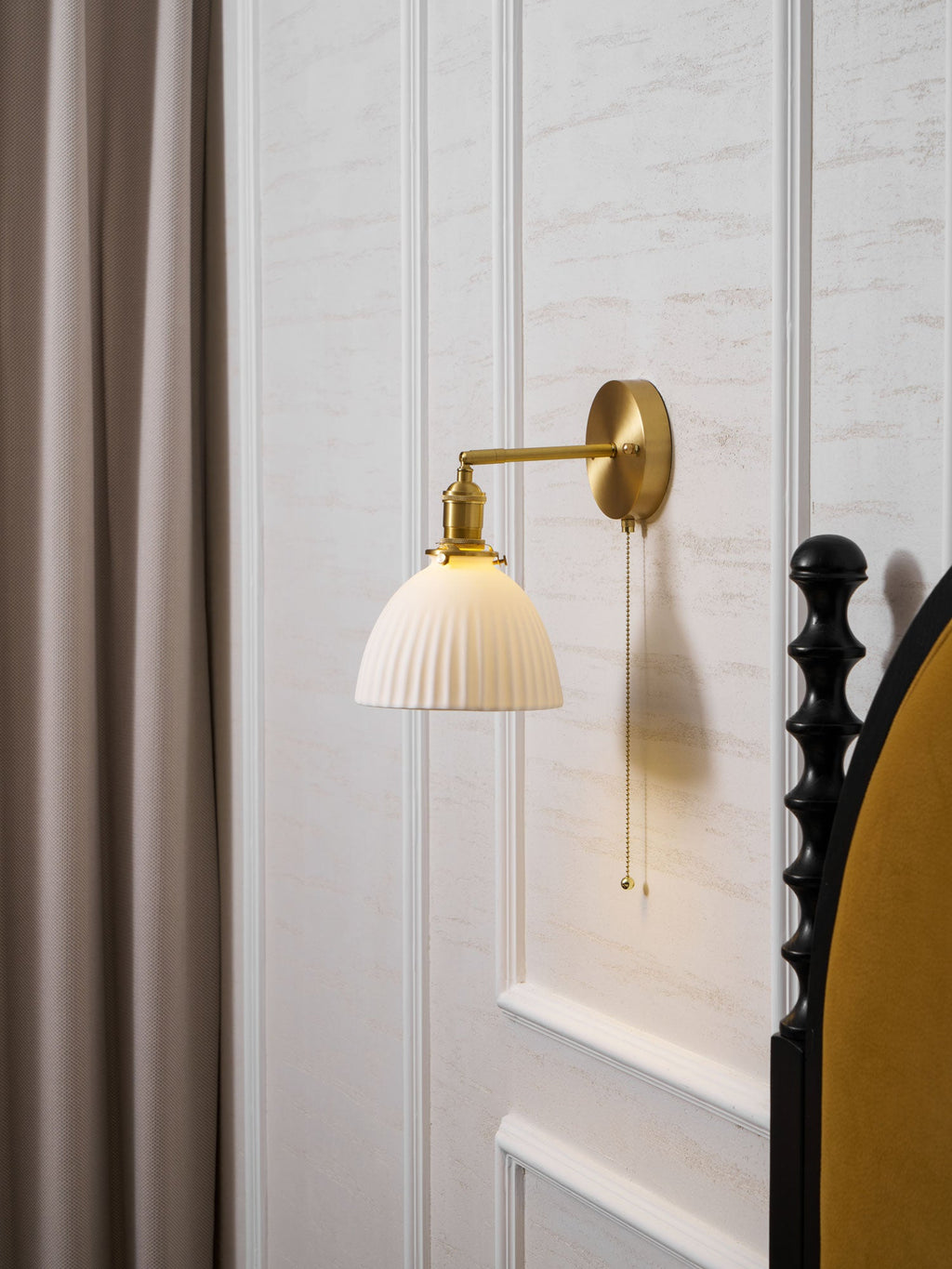This Clérachic wall light hangs on a panelled wall beside a taupe curtain and a mustard velvet headboard.