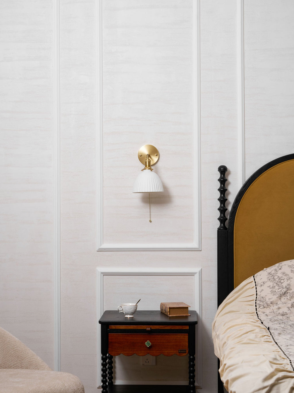 Wider bedroom view of a small Clérachic wall light on a panelled wall, above a dark table with a drawer and a cup.