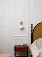 Wider bedroom view of a small Clérachic wall light on a panelled wall, above a dark table with a drawer and a cup.