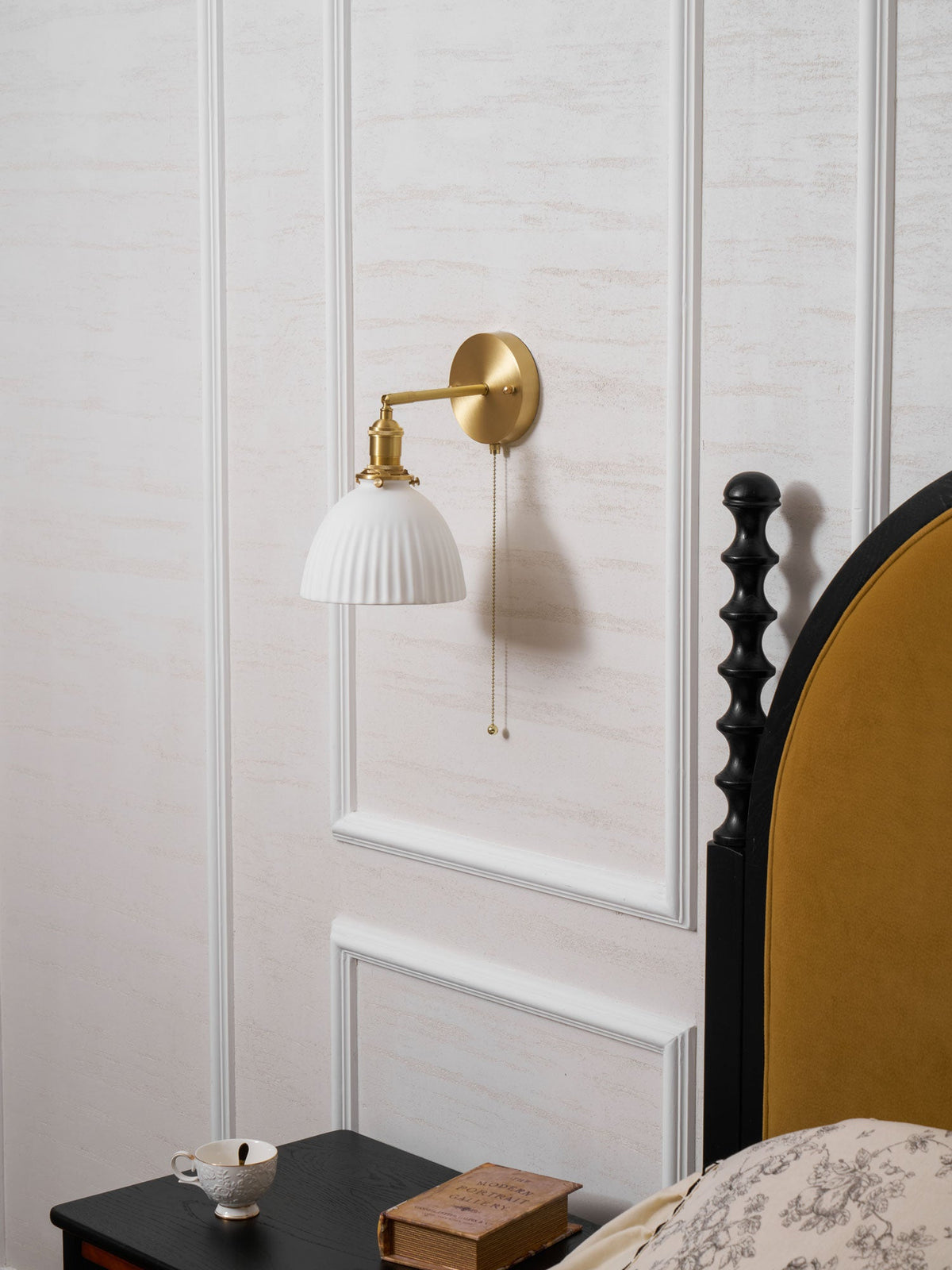 Unlit Clérachic wall light with a pleated shade and brass arm, mounted above a bedside table next to a mustard headboard.
