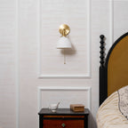 Clérachic wall light with a white ribbed shade glowing faintly above a dark bedside table, beside a curved headboard.