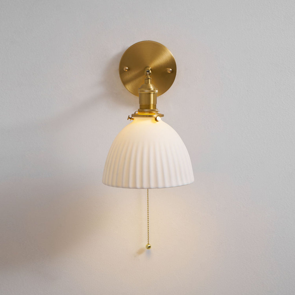 Front view of a lit Clérachic wall light on a grey wall, the round brass backplate above the pleated ceramic shade and pull chain.