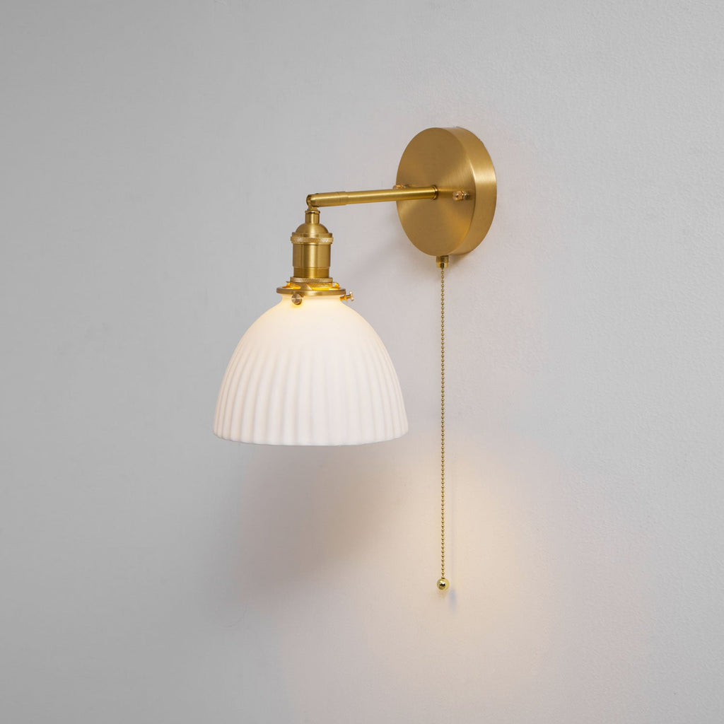 Clérachic wall light with a round brass backplate, a swing arm and a pleated white shade on a light grey wall.