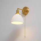 Lit pleated white shade of this Clérachic wall light seen from below, a brass arm and long pull chain over a grey wall.