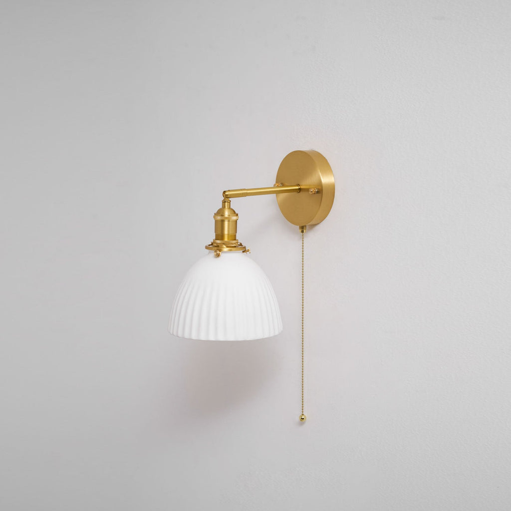 A smaller view of the Clérachic wall light on a grey wall, its brass arm reaching out and a thin pull chain hanging.