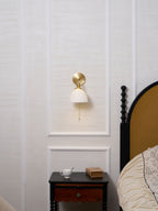 Small Clérachic wall light glowing on a panelled wall above a dark nightstand with a cup and a book, a bed to one side.