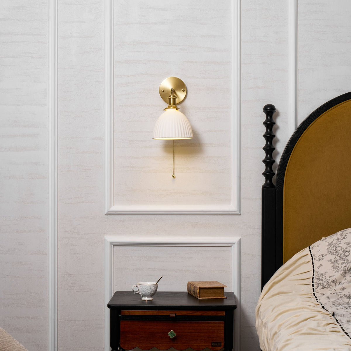 Lit Clérachic brass wall light on a panelled wall, a warm glow beneath the pleated shade, with a mustard bedhead at the side.