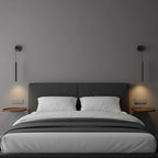 A pair of slim black wall lights flank a grey upholstered bed, each throwing light onto a round wooden shelf, from Clérachic.