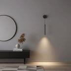 Black tube wall light on a grey wall beside a large round mirror, with a warm pool of light on a low black sideboard, from Clérachic.