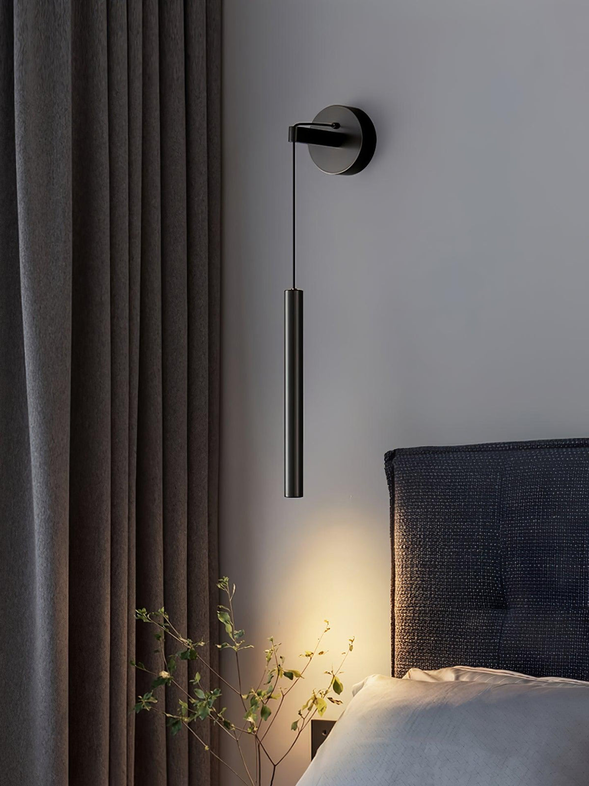 Black wall light on a pale wall beside a heavy grey curtain and a woven headboard, with a warm glow below, from Clérachic.